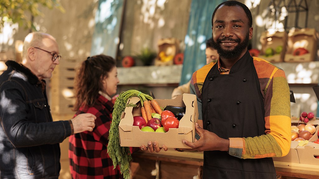 Farmers market seller holding box of fruits and veggies, presenting natural fresh products to clients.
