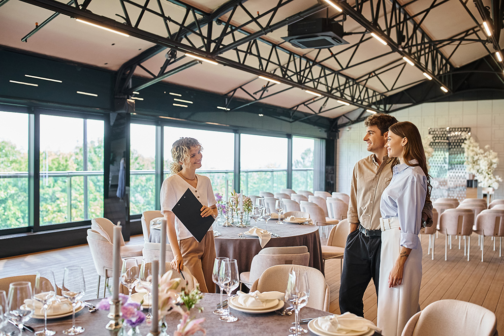 smiling couple talking to event organiser near festive tables in modern wedding venue
