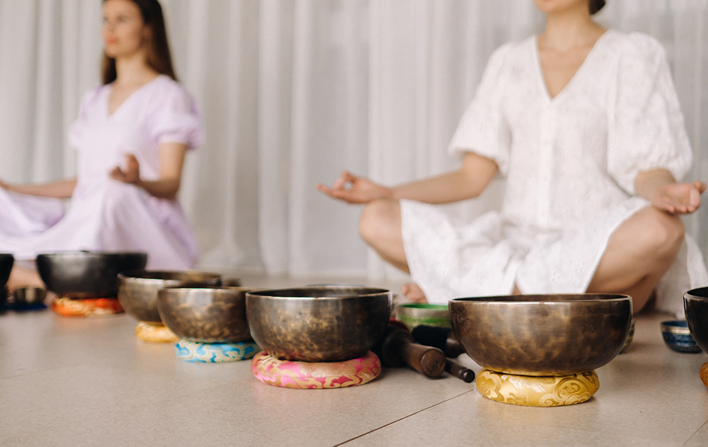 Two women are sitting with Tibetan bowls in the lotus position before a yoga class in the gym.