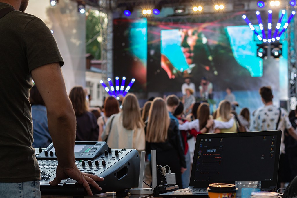 AV technician, laptop outdoors at party festival with stage and crowd of people in background.