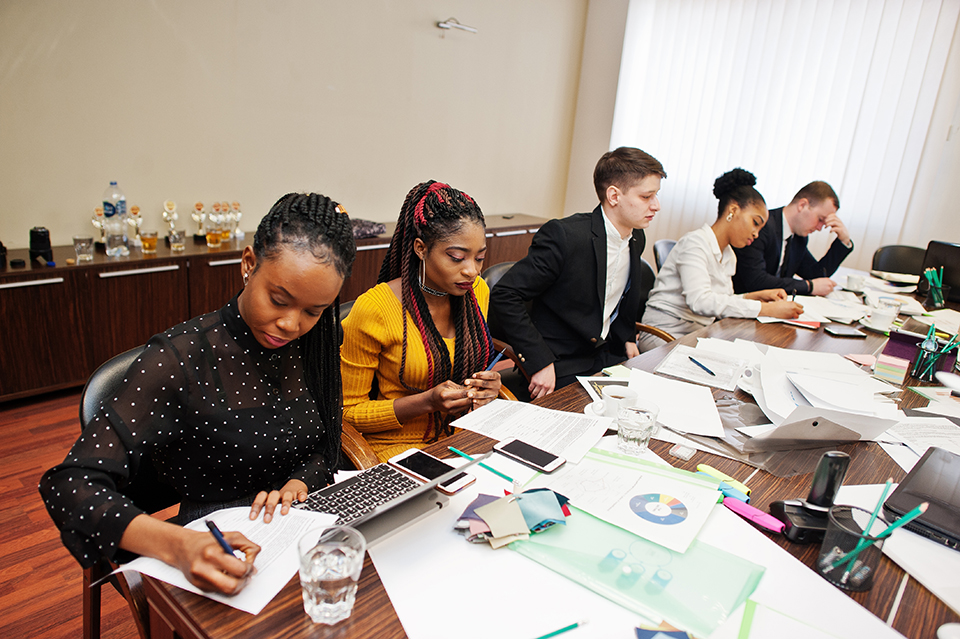 Multiracial business team addressing meeting around boardroom table, working together and write something on papers.