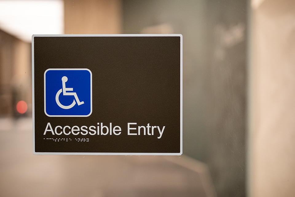 The image shows a sign on a glass door highlighting an accessible entry point with a wheelchair symbol. The background is softly blurred, creating a calm and inclusive atmosphere for a diverse and multi-generational event.